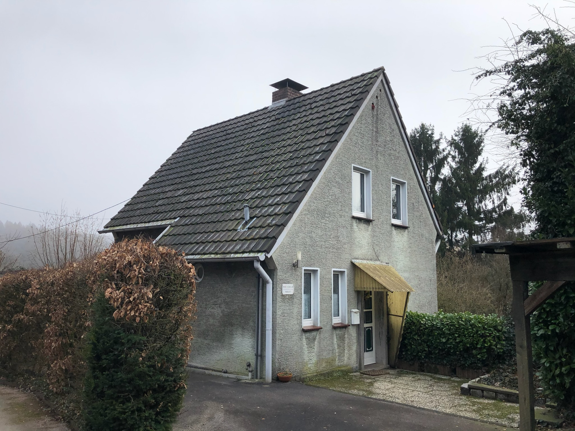 Bestandshaus - Projekt H9 Einfamilienhaus mit grauer Fassade und Satteldach, umgeben von Bäumen und Sträuchern.